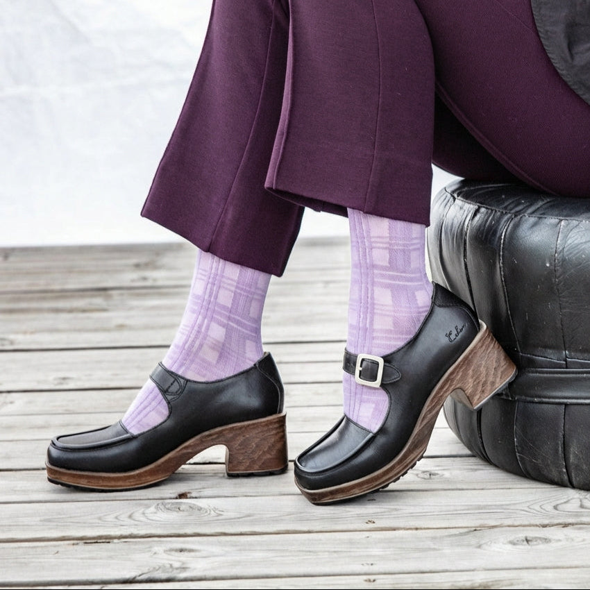 Person wearing black Mary Jane flexible soled clogs, purple pants, sitting on a wooden deck