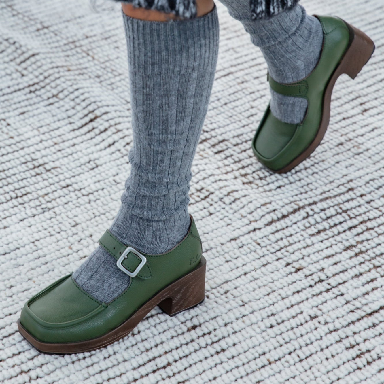 Green loafers with brown soles worn with gray socks on a textured carpet.